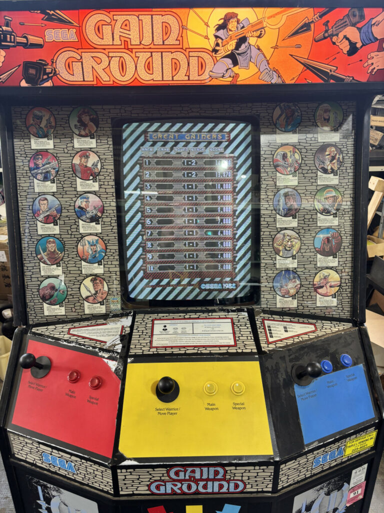 Inquadratura frontale ravvicinata della plancia di comando a tre giocatori del cabinato Gain Ground, completa di joystick e pulsanti originali.