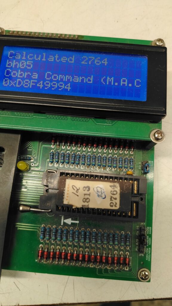 Schermo LCD di un tester diagnostico per EPROM nel laboratorio Arcade Story. L'apparecchio analizza un chip con etichetta originale 12 / 1813 / 2764, rivelando a sorpresa che il contenuto reale appartiene al gioco Cobra Command (M.A.C.) anziché a Super Zaxxon.